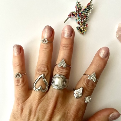 Mão com anéis de prata variados e unhas rosa cintilante, broche de pássaro colorido e flor branca ao fundo