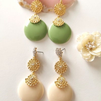 Dois pares de brincos pendentes em dourado com discos em verde e bege, sobre fundo branco com pedra rosa e flor creme