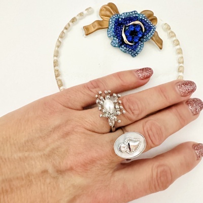 Anel prateado com pérola usado em dedo com unhas com glitter com prato decorativo azul e dourado ao fundo.