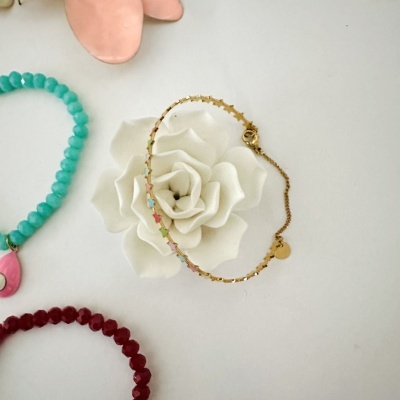 Três pulseiras: dourada com estrelas coloridas, contas turquesa com pendente rosa, contas vermelhas, sobre fundo branco com flor decorativa.