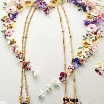 Dois colares dourados com pingentes coloridos sobre fundo branco com flores pastel em forma de coração.