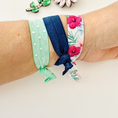 Três pulseiras de tecido coloridas no pulso e um broche flor decorativo ao fundo