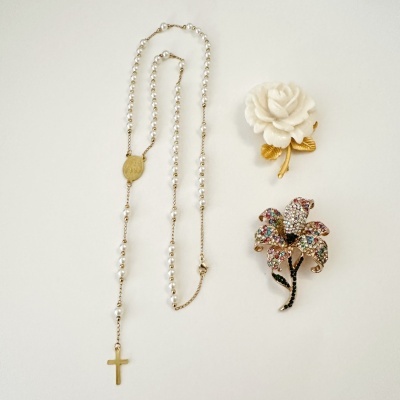 colar de contas brancas com cruz dourada e dois broches florais coloridos sobre fundo branco