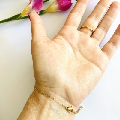 Pulso com pulseira fina dourada, anel e flores coloridas ao fundo