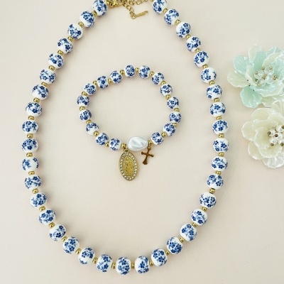 Conjunto de colar e pulseira com contas brancas e azuis e pendentes dourados sobre fundo claro.