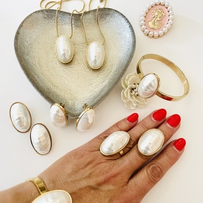 Conjunto de joias douradas com pérolas ovais em bandeja coração e mão com unhas vermelhas a usar anéis