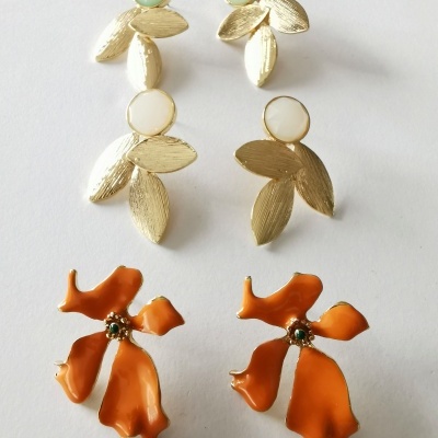 Três pares de brincos: dourados com pedras verdes e branco-opaco, e flores laranja com texturas onduladas