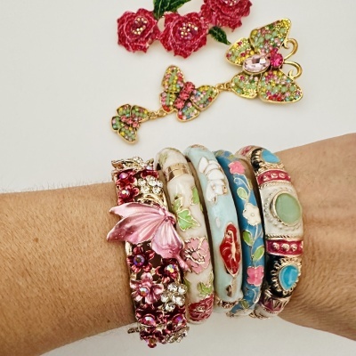 Seis pulseiras coloridas com flores e borboletas em metal dourado com esmalte e pedras coloridas, numa mão. Borboleta dourada decorada acima.