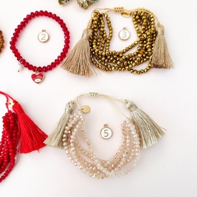 Pulseiras de pedras e contas em vermelho, dourado e pérola clara com franjas, sobre fundo branco.