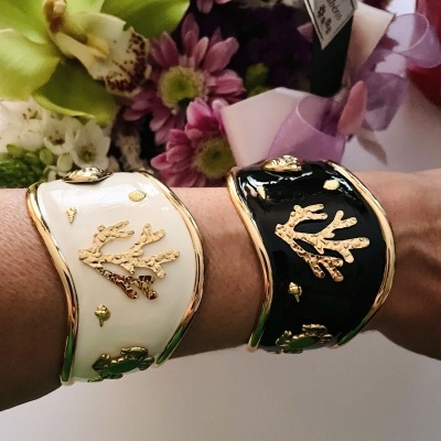Duas pulseiras esmaltadas branca e preta com detalhes dourados num braço, flores e embalagem ao fundo