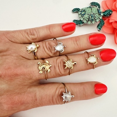 Mão com anéis de tartaruga de diferentes metais e pedras ao lado de tartaruga decorativa verde e flor coral