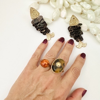 Mão com anéis dourados e pedras, brincos em forma de peixe e adornos perolados