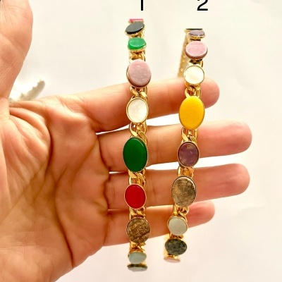 Dois braceletes dourados com pedras coloridas numerados 1 e 2, segurados por uma mão