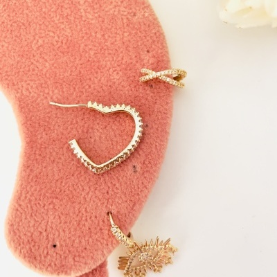 Três brincos dourados com detalhes brilhantes em suporte rosa e fundo branco com flor.