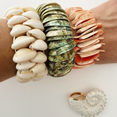 Pulseiras de conchas naturais em várias cores no pulso e anel dourado com concha ao fundo