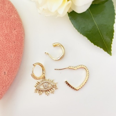 Conjunto de brincos dourados com design de olho e argolas sobre superfície branca com flor e folhas verdes