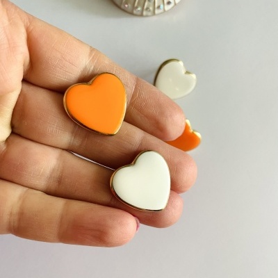 Brincos coração laranja e branco com bordas douradas na mão e superfície branca.