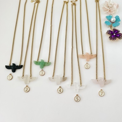 Sete colares dourados com pendentes em forma de passarinho coloridos e medalhas numeradas, sobre fundo branco.