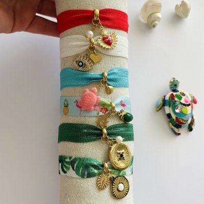 Quatro pulseiras elásticas coloridas com pendentes dourados expostas numa base de tecido bege