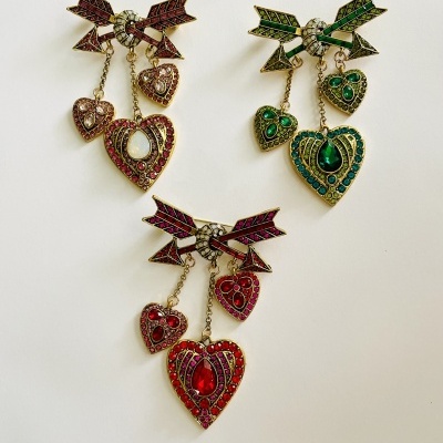 Três broches de coração com flechas em cores vermelho, verde e dourado.