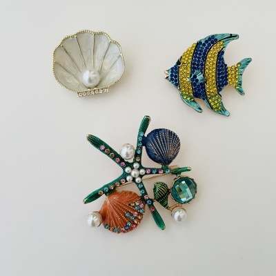 Três broches temáticos marinhos com conchas, peixe e estrela do mar decorados com pérolas e strass