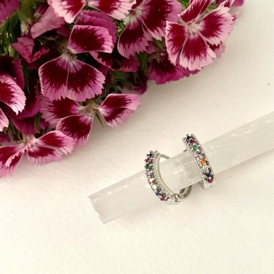 Brincos de argola com pedras coloridas sobre barra translúcida com flores rosas ao fundo