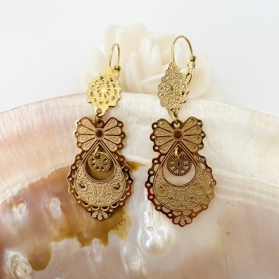 Brincos dourados com padrão floral e forma de laço sobre madrepérola