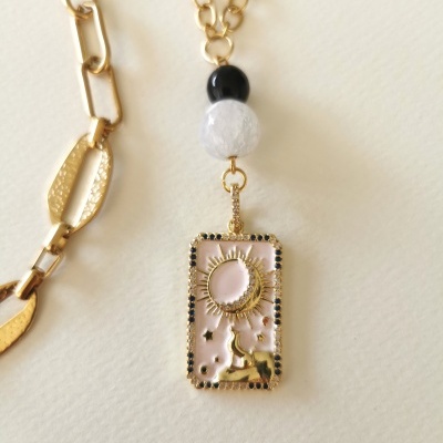 Colar dourado com pendente retangular rosa decorado com lua, estrelas e pedras pretas.