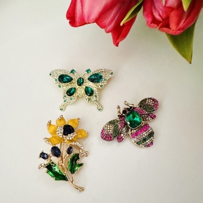 Três broches decorativos coloridos sobre fundo branco com flores cor-de-rosa no canto