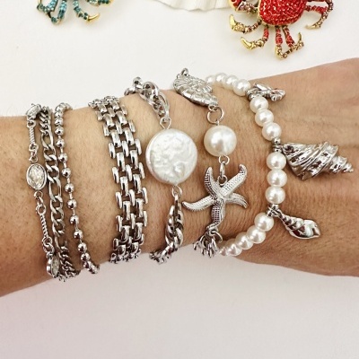 Pulseiras de metal prateado e pérolas com pendentes de estrelas do mar e conchas num pulso com dois broches de caranguejo azul e vermelho ao fundo