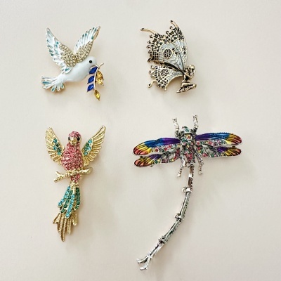 Coleção de quatro pins com desenhos de pombo, fada, pássaro e libélula em diversas cores e materiais.
