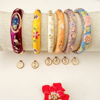 Seis pulseiras esmaltadas com flores em relevo e medalhas numeradas, apoiadas em suporte creme, com flor vermelha ao lado