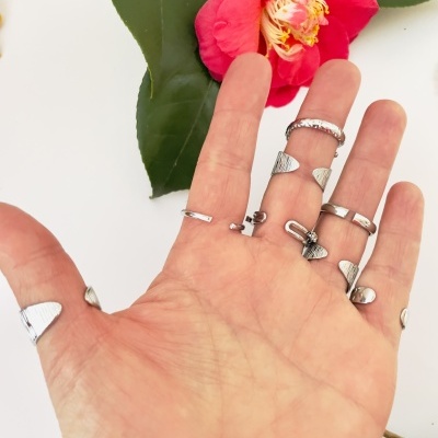 Mão com vários anéis prateados e flor vermelha com folhas verdes ao fundo