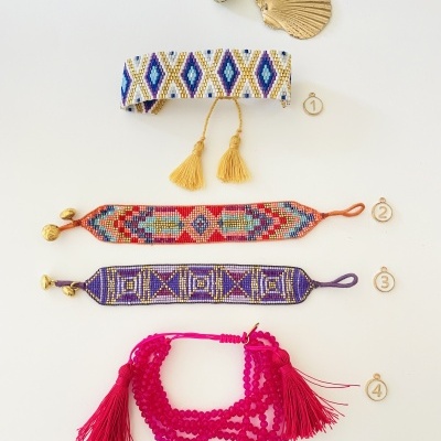 Quatro pulseiras artesanais coloridas com padrões geométricos e franjas sobre fundo branco e duas conchas decorativas