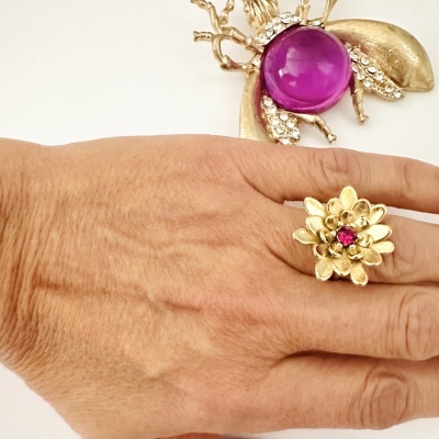 Anel de flor dourado com pedra rosa numa mão e broche de abelha dourado com pedra roxa ao fundo.