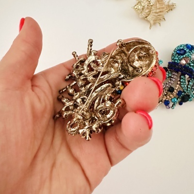 Broche dourado vintage ornamentado na mão com fundo branco, outro broche colorido e concha ao fundo