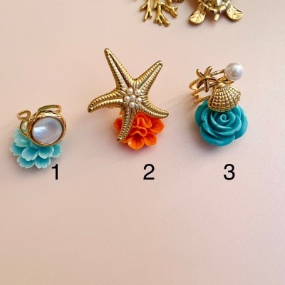 Anéis dourados com flores coloridas e tema marítimo em fundo bege.