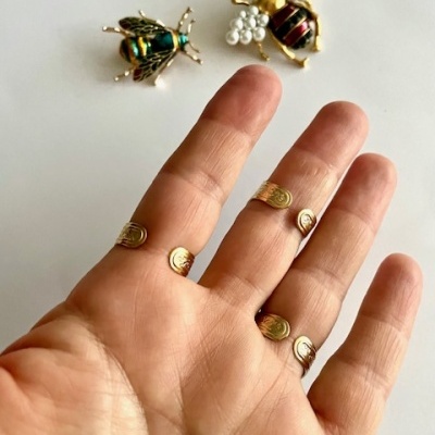 Broches em forma de abelha e inseto metálico com mão ao fundo e anéis dourados