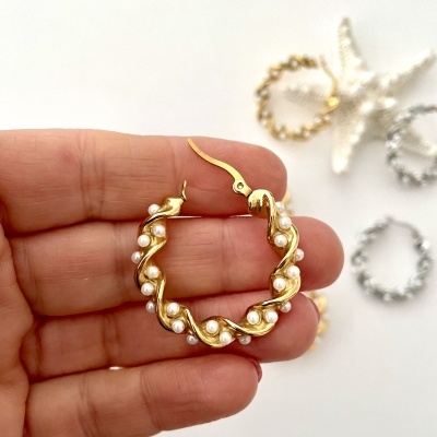 Brincos de argola dourados com pérolas sendo segurados por mão com fundo branco