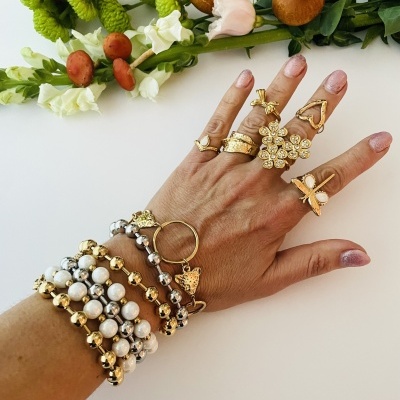 Mão feminina com pulseiras e anéis dourados com flores e cogumelos ao fundo