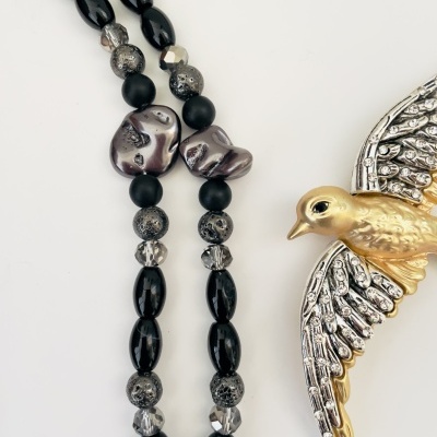 Colar com contas pretas, cinzentas e transparentes e broche de pássaro dourado e prateado.