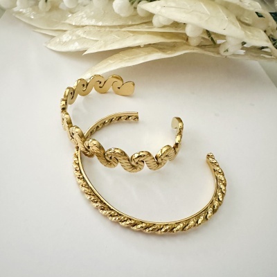 Dois braceletes dourados com padrões ondulados e torcidos sobre fundo branco com flores secas