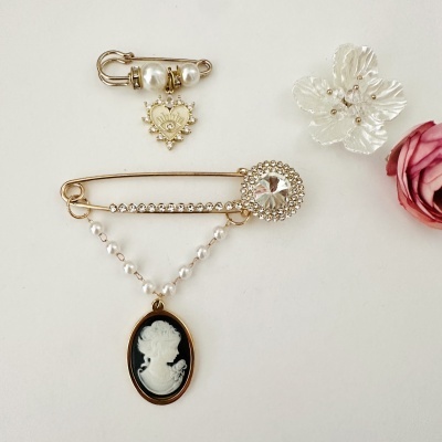 Três alfinetes dourados decorativos, um com pérolas e coração, outro com pedras e medalhão camafeu, e flor acrílica branca.