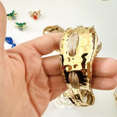 Pulseira dourada com textura martelada e fita entrelaçada, mostrada na mão, com pequenos adornos no fundo branco