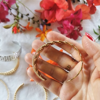 Pulseira dourada texturizada segurada por mão, com pulseiras e flores ao fundo