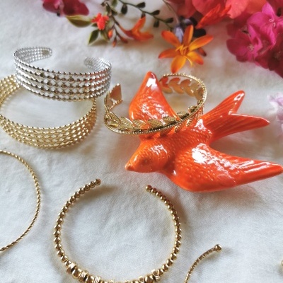 Pulseiras metálicas douradas e prateadas, pássaro laranja decorativo e flores coloridas ao fundo