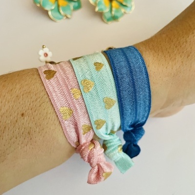 Pulseiras de tecido rosa, azul claro e azul escuro no pulso com brincos de flor ao fundo