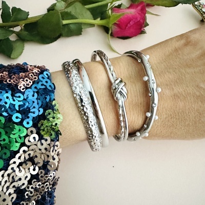 Quatro pulseiras prateadas de diferentes estilos no pulso com rosa ao fundo