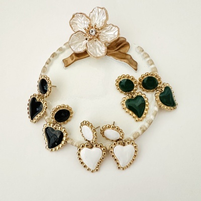 Três pares de brincos em forma de coração com pedras nas cores preto, branco e verde, com acabamento dourado, dispostos sobre suporte redondo claro com flor decorativa.
