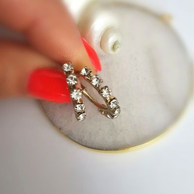 Brinco com pedras brilhantes segurado por mão com unhas vermelhas sobre superfície branca redonda com borda dourada e concha branca.